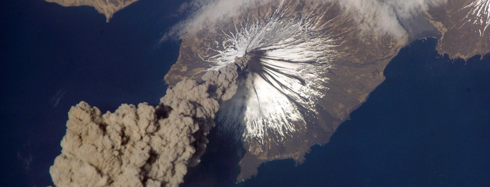 View from space of a volcanic eruption. A gray cloud of ash seen in the bottom left corner is floating away from the volcano site.