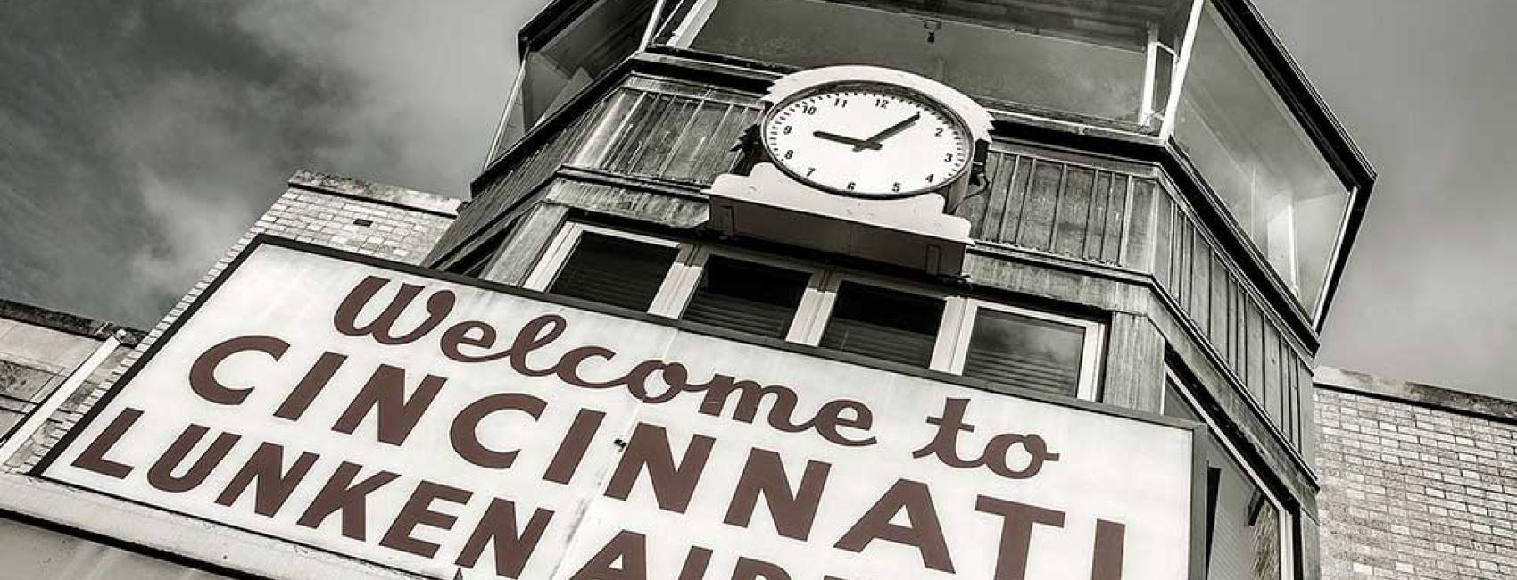 Front view of an air traffic control tower at Cincinnati Municipal Airport. The base is mostly not visible but a sign stating "Welcome to Cincinnati Lunken Airport" is placed under the windows. Above the windows, a clock is visible.