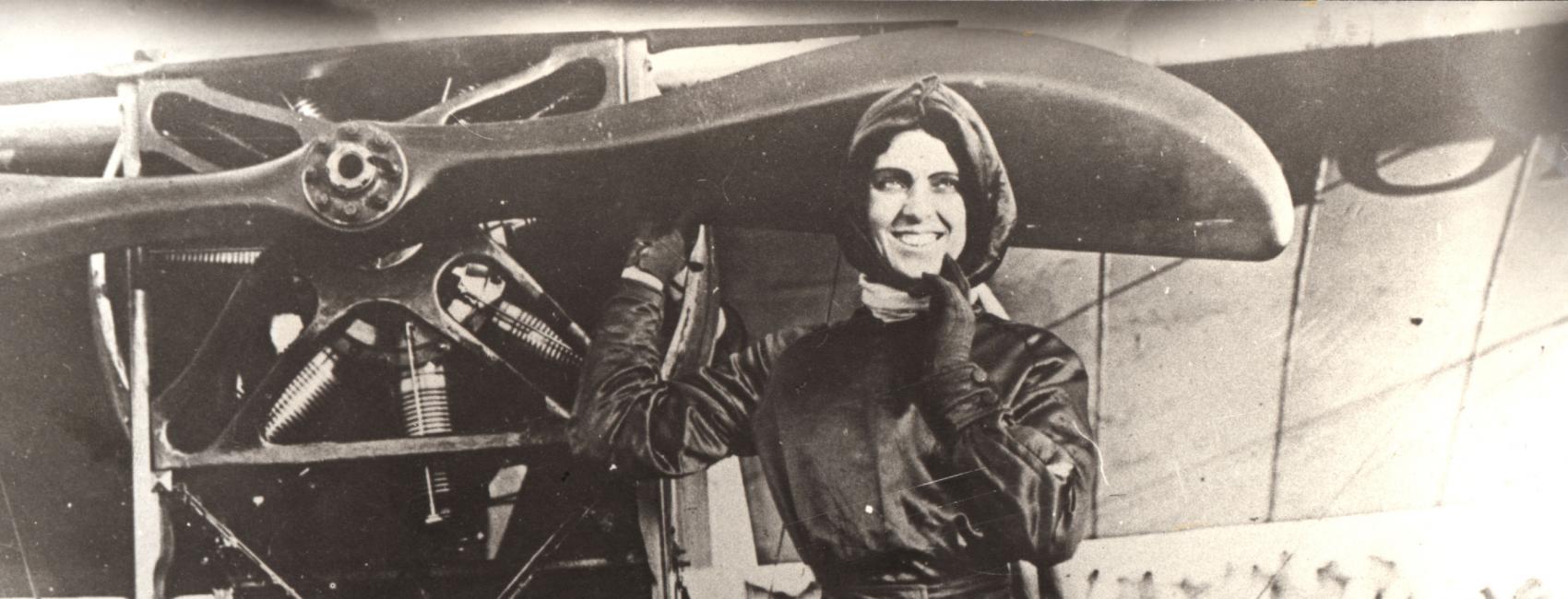 Woman in flying gear poses in front of an airplane. Right hand on propeller, left hand on her chin. Crowd in background.