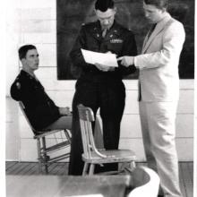 One man sits in a chair while two men stand up and look at a paper. 