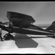 Black and white left side view of monoplane painted black with white stripe down middle. Cursive handwritten text in center of plane reads: "Abyssinia Emperor Haile Salassi I King of Kings." Tail text "782W/Bellanca" Black man by nose right hand in pocket