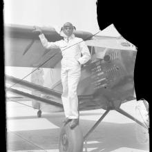 Three quarter right view of monoplane. Black man in white flight suit, poses standing on the right front wheel, holding wing in right hand.  Text under window reads: "Holder World’s Non-Refueling Endurance Record 84 Hrs 33 Mins" cracked upper right corner