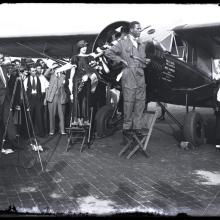 Black and white photo of Black man in flight suit standing on folding chair in front of left side of monoplane. Woman stands behind him holding champagne bottle. Crowd in the background with camera on tripod to the left.