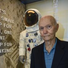 Man in blue shirt stands in front of exhibit case with spacesuit inside.