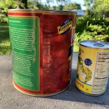 Two cans sit on table. (left) Large 3 quart can of tomatoes with red label (right) Smaller 15.5 ounce can of artichoke hearts with yellow label