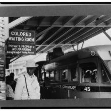 A Black man stands next to a bus and a sign that reads "Colored Waiting Room"