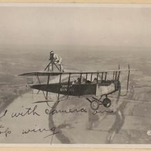 A man performing a barnstorming act standing on the wing of an airplane holding a camera