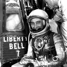 man in silver spacesuit smiles in front of space capsule that says LIBERTY BELL 7