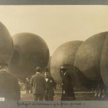 Black and white photo. In the center are three men dressed in suits, the two in the right wearing derbys, the middle man in a top hat. Five hot air balloons in the background. The back of a man in a suit and derby is in the foreground in the lower left corner.