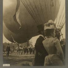 Black and white photo of the backs of a man in a suit and top hat and a woman in a white dress with floral hat with hot air balloons in the background