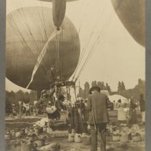 Black and white photograph of a man's back. We can presume that he is using a camera to take a photograph of the three men in the basket of a hot air balloon--the man on the left is partially hanging out of the basket on a rope.