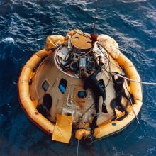 A color photograph of navy divers recovering the Apollo 6 Command Module from the Pacific Ocean. The Command Module floats on the surface of the water.