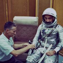 man in silver spacesuit with another man assists him with his glove