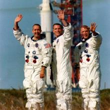 A photograph of the three men who make up the Apollo 9 crew in front of a rocket. All are waving to the camera.