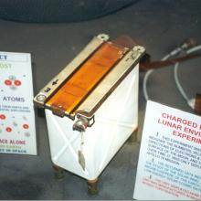 Photograph of the charged particle lunar environment experiment, which is shaped like a small rectangular box that is mostly white with orange on top. Two placards are next to the experiment to explain its function. 