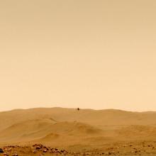 A view of the orange surface of Mars with hills in the background and a barely visible device flying above in the air.