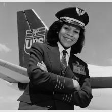 Black & white photograph. African-American woman with hair to shoulders turned to her right with left hand crossed under right.  She is wearing a dark uniform with three pale thin stripes above each hand and an ID badge on her left chest.  She has a white collared shirt with a dark tie with thin pale stripes under the hack. She wears a stiff pilots cap with two laurel leaves on the brim and a star surrounded by wings on the crest.  In the background left is an airplane tail with the UPS logo of wrapped pack