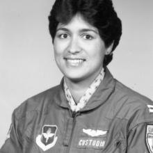 Black and white portrait of woman with short wavy hair, parted in the middle.  Stud earring in left ear (right ear is not visible).  She is wearing a dark jacket with a zipper with a cloth pull.  Patch on her right side (the left of the image) features a icon with wings.  Two patches on her left side (right of photo). Top is wings. Bottom is a nameplate that says "Custodio."  There are two pens in her left sleeve. Another patch is cut off by the edge. White collar with a dark airplane print under the jacket