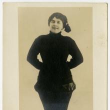 A woman in a jaunty hat stands with her hands behind her back. 