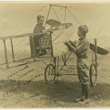 Black and white photograph. One-half right rear view of a man sitting in the cockpit of a monoplane, wearing a helmet with goggles pushed up. To his right a man stands on the ground (back mostly turned) with his hands up in the air. He is wearing a cap.