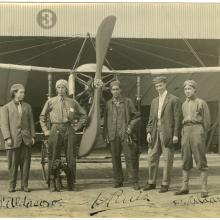 Five men stand in front of a monoplane with a two blade propeller. Background: a hanger made of corrugated metal and to the left of center is the number 3 inside a circle.  From left to right: first man in a suit jacket facing slightly to the right with a backwards cap. second man is wearing a backwards cap and does not wear a jacket and has his left hand in his pocket. The third man is on the other side of the propeller is wearing a cap and unbuttoned jacket.  The fifth man stands with his hands behind him