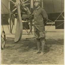 Man stands to the right of a two-bladed propeller with the blades running from the sky to the ground. He is wearing a leather cap with goggles pushed up over the cap and a button down jacket.