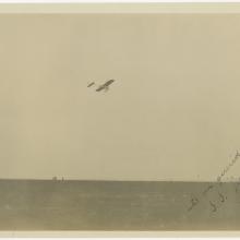 black and white photo One-half right rear view of monoplane in flight with the ground covering the bottom third of the image