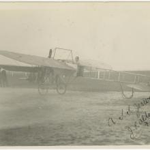 black and white photograph. One-half left rear view of monoplane. man in cap in cockpit. men standing in left background. Number five is marked on the tail of the aircraft