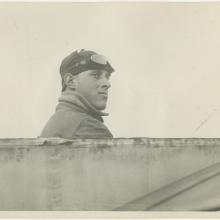 Black and white photo: Right side profile of a man (sitting in a cockpit but you can only see a line across the bottom of the image). Man is wearing a high collared jacket, leather flying cap and goggles pushed up over the cap.