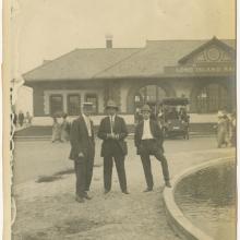 Black and white photo. Three men in suits in foreground. Man on left, turned slightly to his right side is turned to his right and wears a flat brimmed hat.  Man in center wears striped tie and fedora and holds his hands together. Man on right has his left foot up on the edge of a found, wearing a fedora and pale tie. Behind them to the left are two women, one with a parasol. Behind to the right is the back of a car. In background is a building with three rectangular windows on the left and arched windows.