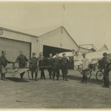 Group of 9 men stand in front of a monoplane. Background: hangars made of corrugated metal. Woman sits in the cockpit with a bulldog.