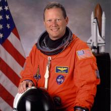 Portrait photograph of man with short sandy brown hair, wearing an orange astronaut jumpsuit with patches (left in shape of shuttle, upper right nameplate "David Brown," bottom right NASA globe logo, American flag on left sleeve.  Right hand resting on helmet.  Blurred background: American flag on left, shuttle model on right.