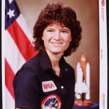 Color photo portrait of a woman with chin length curly brown hair, wearing a navy blue short sleeved polo shirt with two patches. Top patch: NASA name logo.  Bottom patch: Circular mission patch with Challenger Space shuttle and astronaut's name.  Blurred Background: on the left American flag, on the ride Space Shuttle model. Border: NASA photo markings.