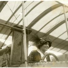 A woman in a hat with three pins and a feather behind a steering wheel.