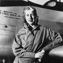 A woman in aviator gear leans against an airplane wing. 