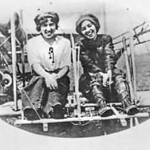 Two sisters sit in an airplane, laughing and smiling.