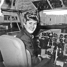 A woman turns around and smiles from the cockpit of a plane.