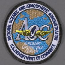 Embroidered National Oceanic and Atmospheric Administration (NOAA) Aircraft Operations Center (AOC) insignia; gold wings in center over white letter text "AOC", "AIRCRAFT OPERATIONS CENTER"; background map of the Atlantic Ocean depicted in blue with tan continental land masses; map surrounded by white band with navy blue letter text "NATIONAL OCEANIC AND ATMOSPHERIC ADMINISTRATION U.S. DEPARTMENT OF COMMERCE"; black edge.