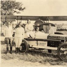 Five people lounge on a car labeled "Ruth Law's Flying Circus." Behind the car is an airplane.