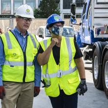 Two people stand with safety vest and hardhats on outside.