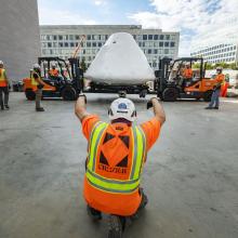 Apollo 11 command module Columbia being lifted by two forklifts and surrounded by workers.