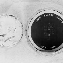 A black and white photograph of a circular disc with "from planet Earth, July 1969" written in large font. Smaller type is in the middle of the disc, but is not legible. The disc is next to a quarter for scale, and is roughly the same size.