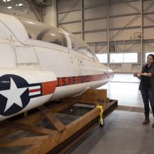 Two women examine a T-38 Talon aircraft while it's strapped to the back of a truck without its landing gears.