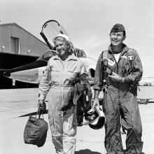 Black and white image of Jackie Cochran and Chuck Yeager walking and smiling with an aircraft in the background.