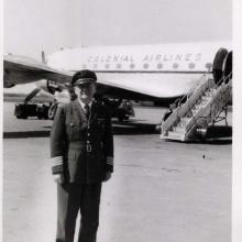 Black and white photograph. Foreground: Man in dark pilot's uniform with a cap and stripes at the ends of his sleaves. Background: Side of a passenger airplane labelled "Colonial Airlines" above circular windows. A set of airstairs is behind and to the right of the man.