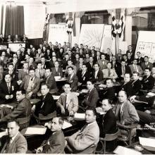 Black and white photograph of a convention of men in a large room, seated at tables. In the background on left is a podium with an eagle symbol on it and an American flag hanging vertically with the stars on the left.