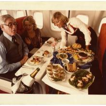 A color photo of a flight attendant serving food to two passengers aboard a plane.