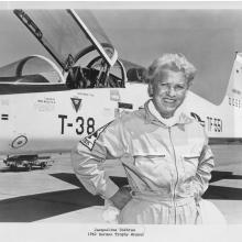 An aged Jackie Cochran posing with her hands on her hips and smiling and a T-38 Talon aircraft in the background.