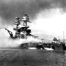 Black and white image of a battleship in flames and smoke as sits on a body of water.