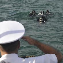 A man in a white military uniform and hat is pictured from behind as he salutes divers in a body of water.